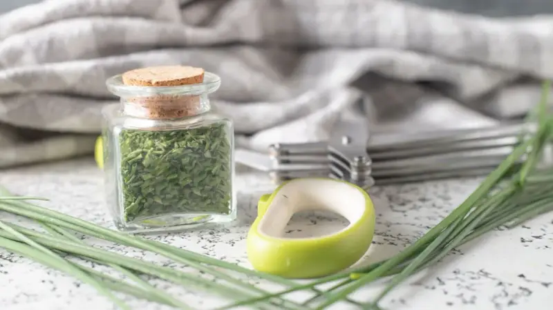 A jar filled with dried chives, showcasing how to dry chives for long-term storage. Fresh chives and kitchen tools are nearby.
